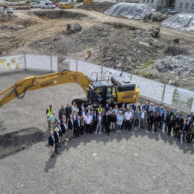 Ein Gruppenfoto aus der Luft aufgenommen. Die Menschen stehen auf einem Baugrundstück, hinter ihnen ist ein Bagger.