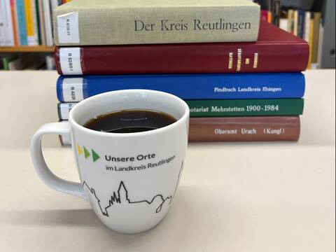 Ein Stapel Bücher liegt im Hintergrund auf einem Tisch vor einem Bücherregal, während im Vordergrund eine Tasse mit Kaffee steht, auf der "Unsere Orte im Landkreis Reutlingen" steht.