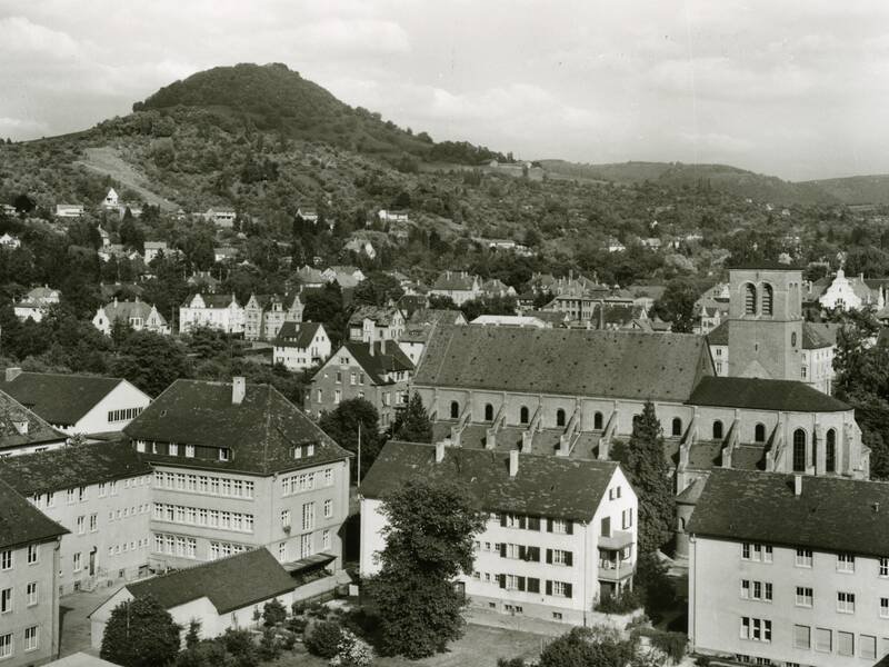 Schwarz-weiß-Fotografie einer Stadt mit traditionellen Gebäuden, einer großen Kirche und einem Hügel im Hintergrund.