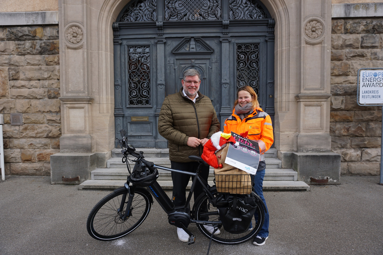 Zwei Personen stehen vor einem historischen Gebäude, eine hält eine Karte und steht neben einem Fahrrad mit einem Korb voller Pakete.