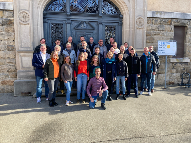 Eine Gruppe von etwa 25 Menschen posiert lächelnd vor einer großen, dekorativen Tür aus Metall und Stein, wobei rechts ein Schild mit der Aufschrift "European Energy Award" sichtbar ist.