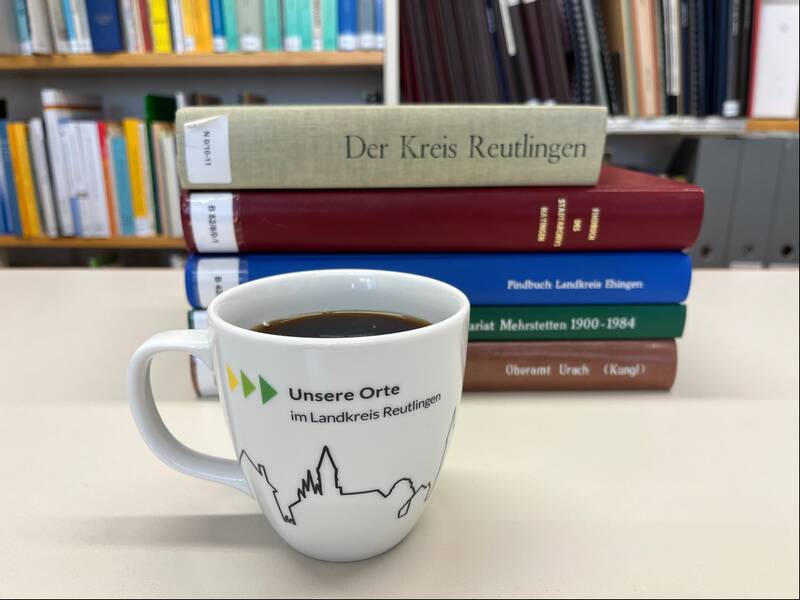 Eine Tasse Kaffee mit der Aufschrift "Unsere Orte im Landkreis Reutlingen" steht vor einem Stapel Bücher, im Hintergrund ein Bücherregal.